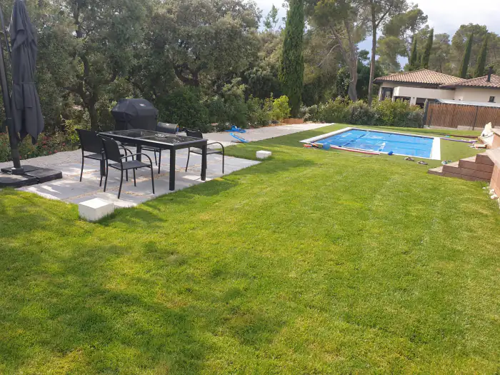 Grande pelouse verte devant une piscine et un coin repas ombragé